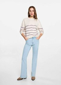 Suéter de punto beige con rayas horizontales moradas, combinado con jeans acampanados de color azul claro y zapatos planos puntiagudos marrones, con las manos en los bolsillos.