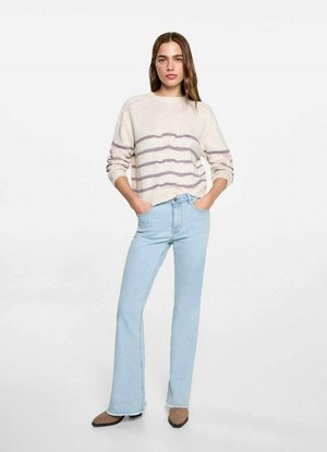Suéter de punto beige con rayas horizontales moradas, combinado con jeans acampanados de color azul claro y zapatos planos puntiagudos marrones, con las manos en los bolsillos.