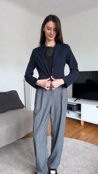 Γυναίκα που κουμπώνει ένα μπλε σκούρο σακάκι πάνω από ένα μαύρο τοπ, φοράει γκρι φαρδιά παντελόνια και στέκεται σε ένα σαλόνι με τηλεόραση και καναπέ.