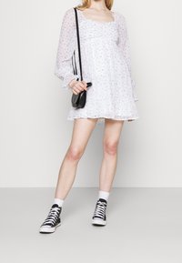 Biała kwiatowa mini sukienka z długimi, bufiastymi rękawami, marszczonym dekoltem i falbanką u dołu. Połączona z czarnymi tenisówkami za kostkę i małą torebką.