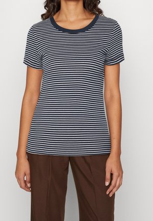 Kobieta ubrana w granatowo-biały, poziomo prążkowany t-shirt z krótkim rękawem i okrągłym dekoltem oraz brązowe spodnie, stojąca na jednolitym tle.