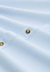 Lichtblauwe stof met een getextureerde afwerking, voorzien van gouden knopen en gestikte naden. Het patroon is subtiel en uniform over het materiaal.