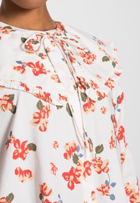 Blusa floral em tecido leve branco com padrões de flores vermelhas e azuis. Apresenta um colarinho franzido, botões à frente e um decote amarrado.