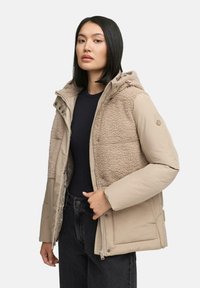 Beige gewatteerde jas met een pluche, bontachtige voorkant. Voorzien van een capuchon, ritssluiting en zijzakken. Gestyled met donkerblauwe jeans en een marineblauw top.