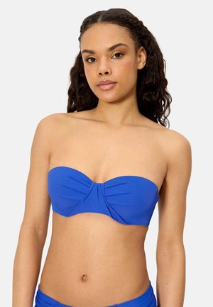 Mujer joven con cabello rizado, vistiendo un top de bikini azul sin tirantes, de pie contra un fondo claro y liso.