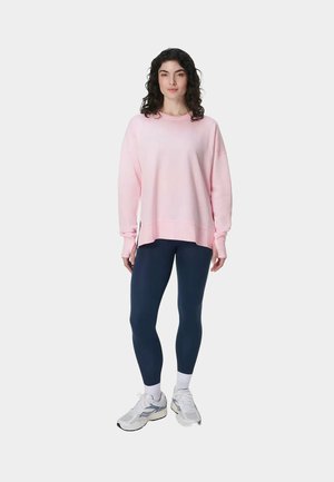 Vrouw staand, gekleed in een loszittende roze sweatshirt, marineblauwe legging, witte sokken en witte hardloopschoenen tegen een effen lichte achtergrond.