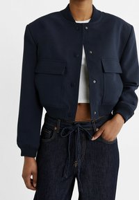 Cropped navy jacket in smooth fabric with ribbed cuffs, snap buttons, and two front pockets, paired with dark denim jeans.