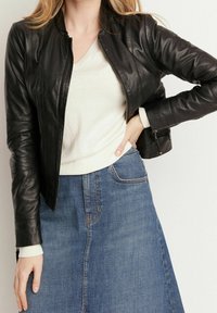 Svart skinnjacka med figursydd design, dragkedja och ribbade detaljer, kombinerad med en kremfärgad tröja och jeanskjol.