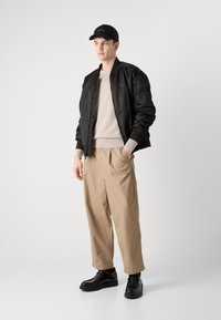 Black bomber jacket over a beige sweater, paired with loose beige trousers and black lace-up boots. Man stands against a neutral background.