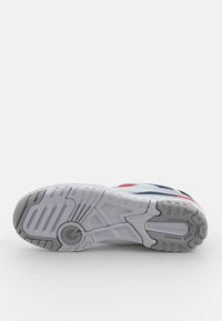 Bílá podešev tenisky s šedým vzorem pro trakci, se červenými a námořnickými akcenty. Logo "New Balance" viditelné na straně. Texturovaný gumový materiál.