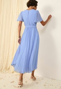 Robe maxi plissée bleu clair avec des manches courtes à volants, taille cintrée et jupe fluide ; portée avec des talons blancs à lanières et des accessoires minimalistes.