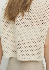 Cremfarbener, kurzer Pullover mit offenem Strickmuster, das ein quadratisches Design und strukturierte Kanten aufweist, getragen über einem hellen Kleidungsstück.