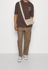 Muž v hnědém tričku s potiskem, khaki cargo kalhotách, teniskách se zebrovaným vzorem a béžové crossbody tašce na pozadí.