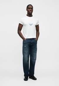 Άνδρας με λευκό κοντομάνικο μπλουζάκι με μπλε λογότυπο, σκούρο μπλε τζιν με ελαφριά φθορά, μαύρα αθλητικά παπούτσια, στέκεται με τα χέρια στις τσέπες.