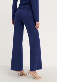 Marine brede pijpenbroeken van een gestructureerde stof, met een hoge taille en subtiele riemdetails. Gecombineerd met doorzichtige haksandalen, die een elegante uitstraling hebben.