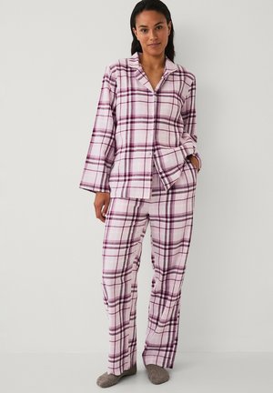 Frau steht und trägt ein rosa-lila kariertes langärmliges Pyjamashirt und passende Hose mit grauen Hausschuhen vor einfachem Hintergrund.