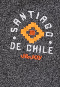 Grijze stof met geborduurde tekst. Bevat "Santiago de Chile" in het wit, een geel-oranje geometrisch ontwerp en "J&Joy" in het oranje.