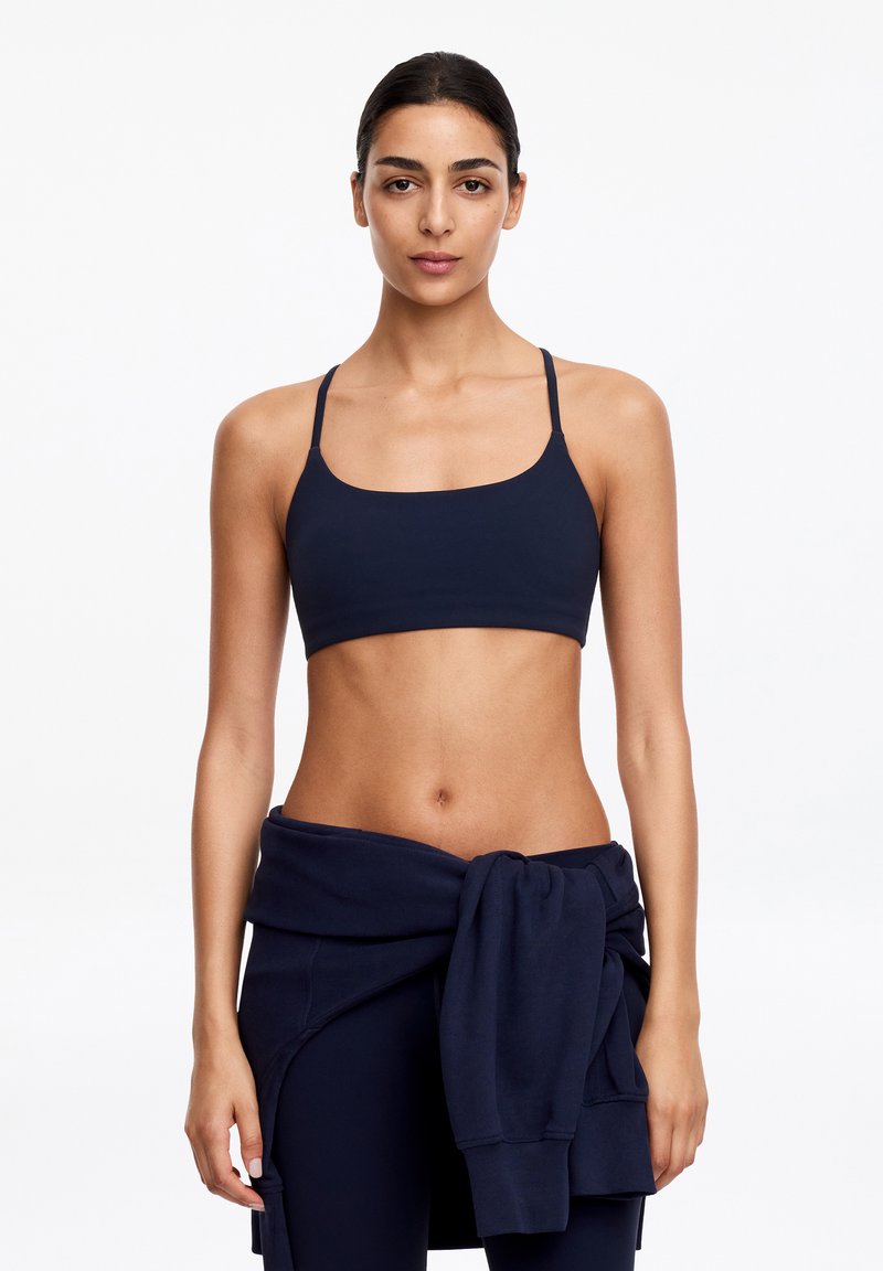 Femme aux cheveux foncés attachés en arrière portant un soutien-gorge de sport bleu marine, des leggings bleu marine et un sweat-shirt bleu marine noué autour de la taille, se tenant devant un fond blanc.