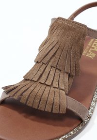 Sandale en daim marron avec un détail de franges superposées sur la bride des orteils et une bordure argentée scintillante le long du bord de la semelle.