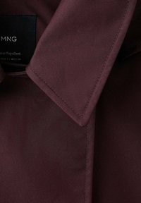 Detalle del cuello de una chaqueta impermeable de color burdeos oscuro con costuras visibles y una etiqueta de marca parcialmente visible "MNG".