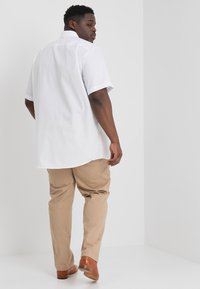 Korte mouw witte shirt met een rechte snit, gecombineerd met lichte beige broek en bruine schoenen, vanuit de achterkant gezien tegen een witte achtergrond.