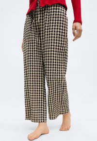 Άτομο που φοράει χαλαρά μαύρα και μπεζ καρό παντελόνια και κόκκινο πλεκτό πουλόβερ, στέκεται ξυπόλητο σε λευκό φόντο.