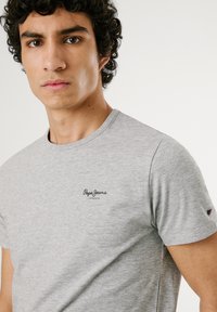 Ung mand med krøllet mørkt hår iført en lysegrå Pepe Jeans London t-shirt mod en ensartet hvid baggrund.