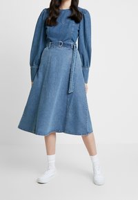 Denim midi-klänning med långa puffärmar, midjebälte, ljusblå färg, slät textur och A-linjeform, med en Chevron-söm vid bröstet.