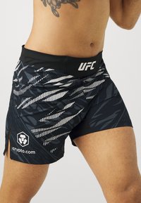 Juodi sportiniai šortai su tekstūruotu grafiniu ra�štu tamsiomis atspalviais. Juosmens juostoje yra UFC logotipas, o šone - crypto.com logotipas.