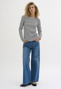 Long-sleeve striped top in black and white, paired with loose-fitting blue jeans. Model wears black flats, hands in pockets, on a plain background.