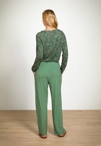 Groene bloemerige top met lange mouwen en een ronde halslijn, gecombineerd met groene gestreepte wijde broek. Platte sandalen zichtbaar aan de voeten.
