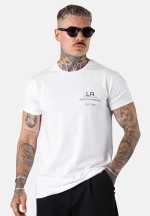 Homme avec des tatouages et une moustache portant des lunettes de soleil noires, un T-shirt blanc imprimé et un pantalon noir, se tenant avec une main dans la poche.