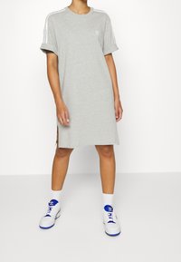 Grå bomullst-shirtklänning med uppvikta ärmar, vita axelränder, sidsprund och en liten logotyp på bröstet. Bärs med vita sneakers.