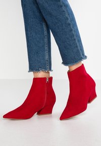 Czerwone zamszowe botki za kostkę z ostrym noskiem, z zamkiem po boku i niskim słupkowym obcasem. Spodnie z falbaniastym wykończeniem nad botkami.