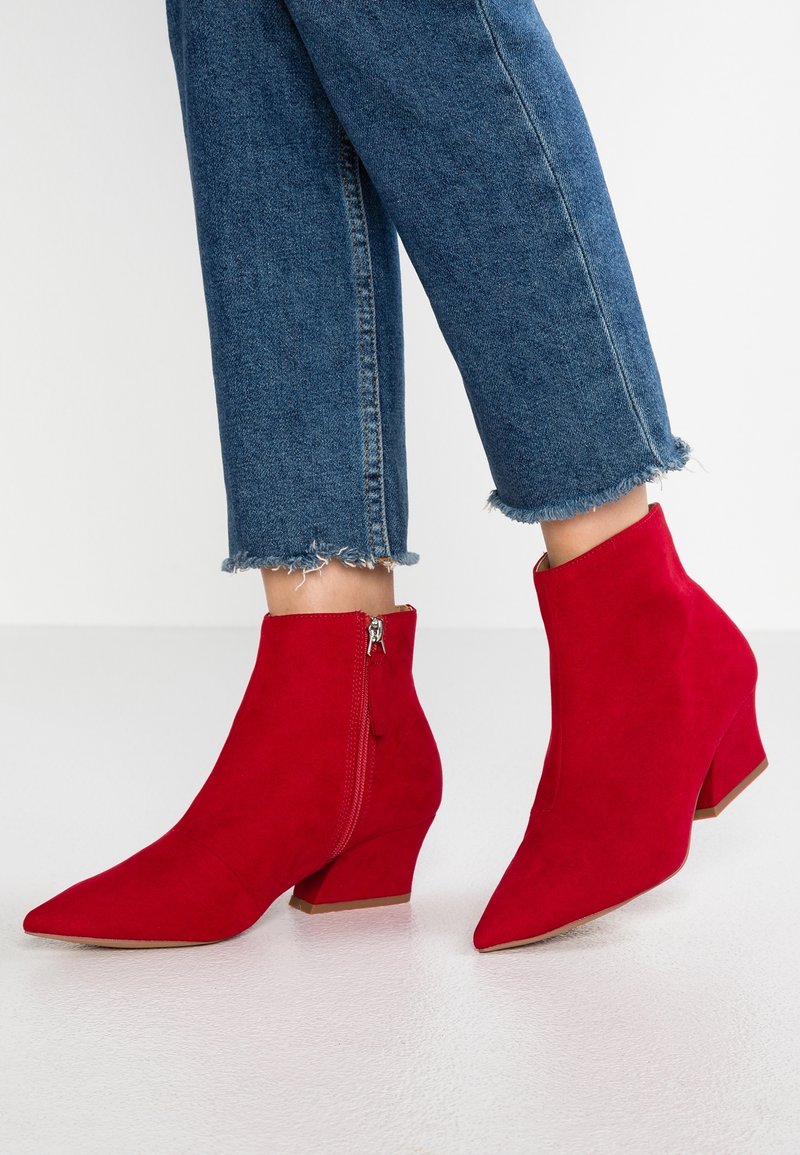 Czerwone zamszowe botki za kostkę z ostrym noskiem, z zamkiem po boku i niskim słupkowym obcasem. Spodnie z falbaniastym wykończeniem nad botkami.