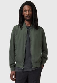 Oliwkowa kurtka bomber z zamkiem błyskawicznym, ryżowymi mankietami i dołem. Posiada subtelne logo na klatce piersiowej oraz gładki, lekki materiał.