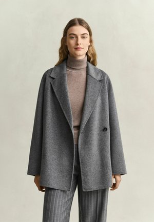 Frau mit langen Haaren trägt einen grauen Wollmantel über einem beigen Rollkragenpullover und grauen Nadelstreifenhosen vor einem einfarbigen hellen Hintergrund.