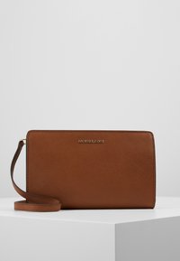 Clutch da polso in pelle marrone con texture, con logo dorato Michael Kors e tracolla in pelle abbinata, poggiata su una superficie bianca.