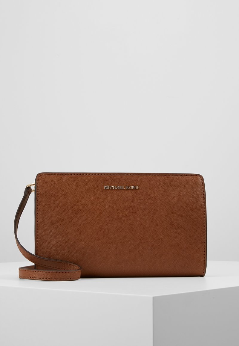 Clutch da polso in pelle marrone con texture, con logo dorato Michael Kors e tracolla in pelle abbinata, poggiata su una superficie bianca.