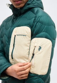 Personne portant une doudoune verte avec des poches zippées beiges sur la manche et la poitrine, main tenant le zip de la manche.