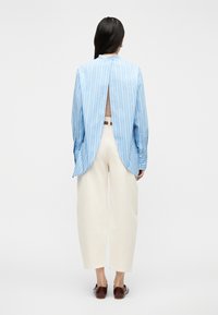 Γυναίκα με μακριά σκούρα μαλλιά φοράει ανοιχτό μπλε ριγέ πουκάμισο με ανοιχτή πλάτη, εκρού παντελόνι με φαρδιά μπατζάκια και σκουρόκαφέ παπούτσια, στέκεται σε λευκό φόντο.