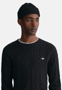 Schwarzer Kabelstrickpullover mit geripptem Rundhalsausschnitt und kleinem Logo auf der Brust. Getragen mit einer passenden schwarzen gerippten Strickmütze.