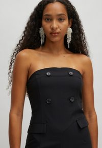 Zwarte strapless jumpsuit met een gestructureerd ontwerp, voorzien van dubbele knopen en zijzakken. Model draagt lange pareloorbellen.