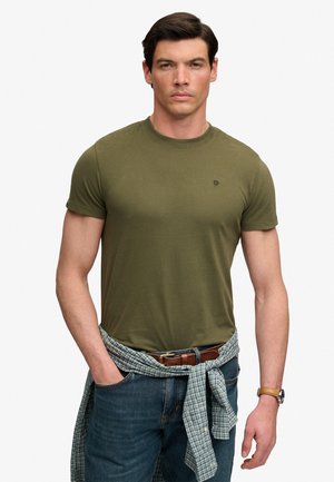 Mężczyzna w oliwkowo-zielonym t-shircie, niebieskich dżinsach, koszuli w kratę związanej wokół talii, brązowym pasku i zegarku na rękę, stojący z jedną ręką w kieszeni.