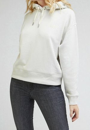 Nainen pukeutuneena vaalean beigeen Lee-huppariin, jossa on kiristysnauhat, ja tummanharmaisiin farkkuihin, yksivärisen taustan edessä, rajattu kaulan ja reisien kohdalta.