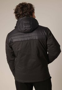 Veste à capuche noire avec une texture matelassée sur les épaules. Présente un bas lisse et mat avec des accents de tissu sombre.