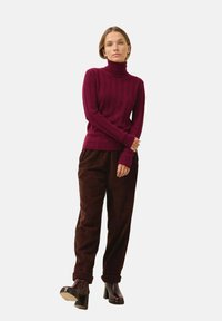 Suéter burdeos de cuello alto acanalado con manga larga, combinado con pantalones de pana marrones; la modelo lleva botas de tacón negras hasta el tobillo.