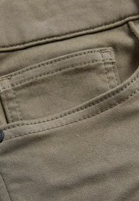 Material textil din bumbac bej, în prim-plan, cu un buzunar ce prezintă detalii vizibile ale cusăturii și o textură netedă.