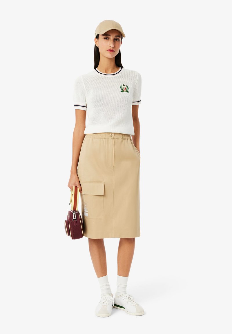 Witte zachttextuur korte mouwen trui met groene borduursels, gecombineerd met een beige knielange rok met een zijzak. Witte sneakers en een pet.