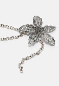 Srebrna ogrlica sa lancem koja sadrži metalni privjesak u obliku lista ukrašenog prozirnim rhinestones i složenim detaljima.